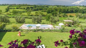 Un espace piscine agrémenté de chaises longues et d’un kiosque, le tout entouré d’une végétation luxuriante et de douces collines.