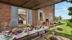 Uma área de refeições ao ar livre com uma casa de tijolo, um alpendre coberto e vista para o campo.