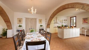Un espace repas avec une longue table dressée pour six personnes, entourée de chaises. On y trouve également une cuisine avec des meubles blancs et un plan de travail.