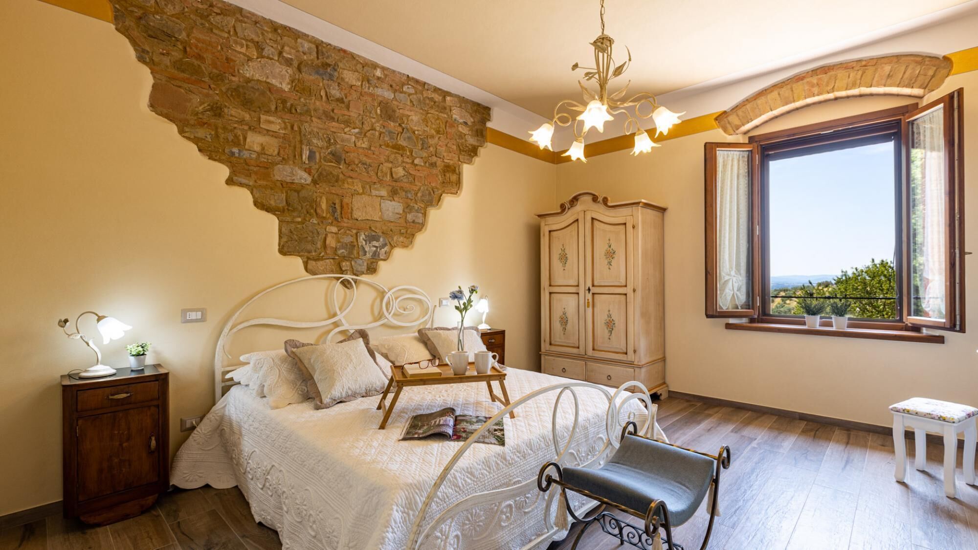 Un dormitorio con pared de piedra, una cama con ropa de cama blanca, una mesita de noche de madera, un aparador y una ventana con vistas.