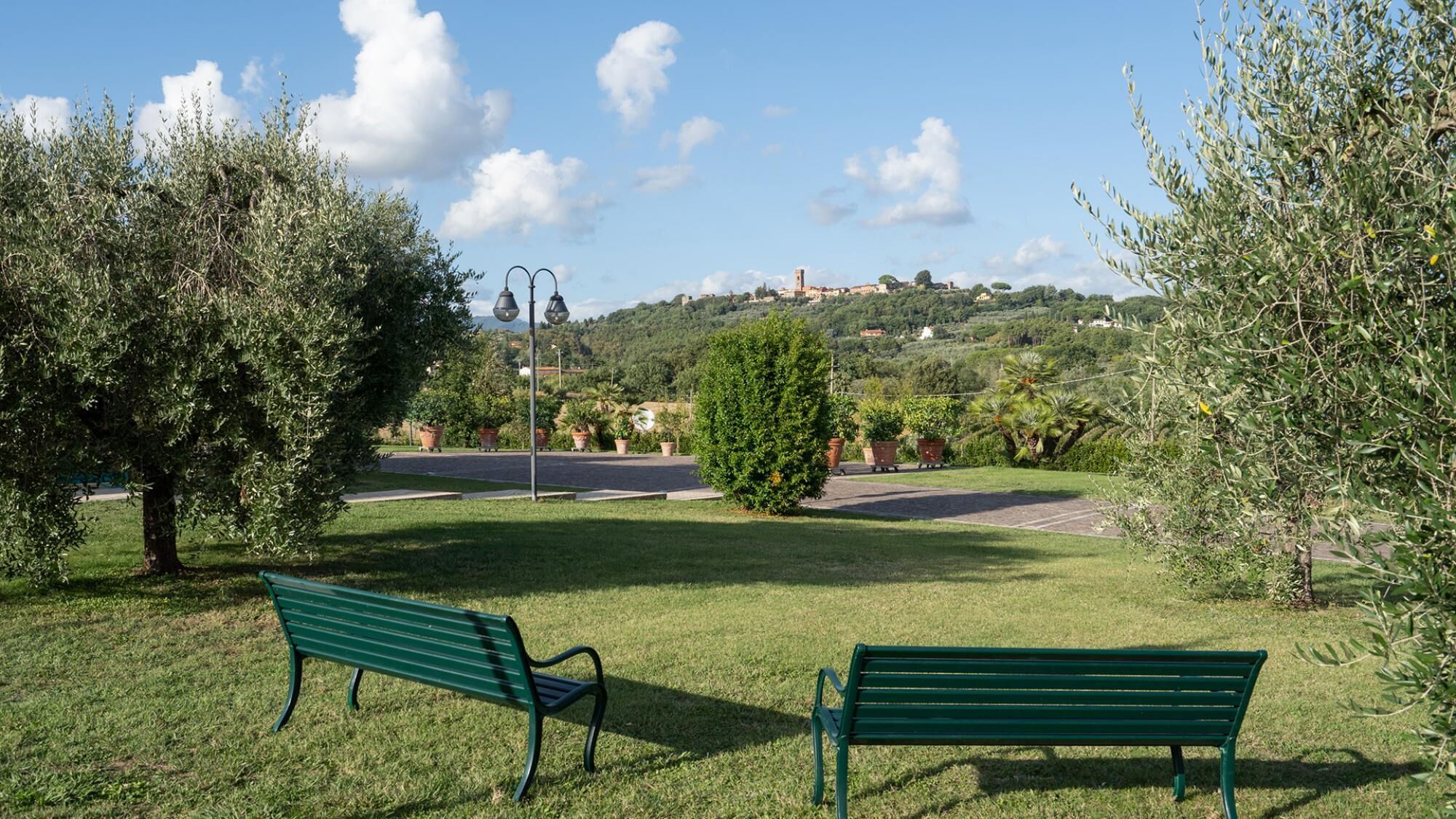 Un parc avec deux bancs verts, une route et une colline sur laquelle se trouvent des bâtiments en arrière-plan.
