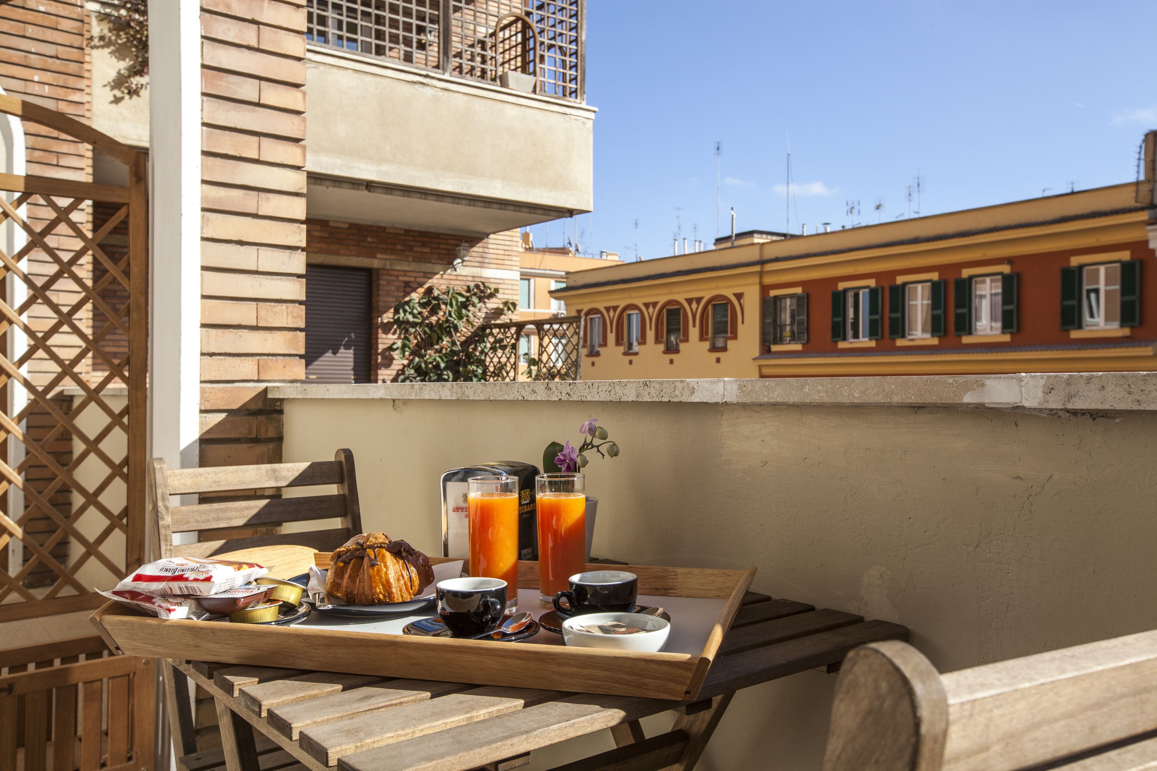Balcón con una bandeja de desayuno que incluye pan, café y jugo, con vista a un paisaje urbano con edificios y cielo despejado.