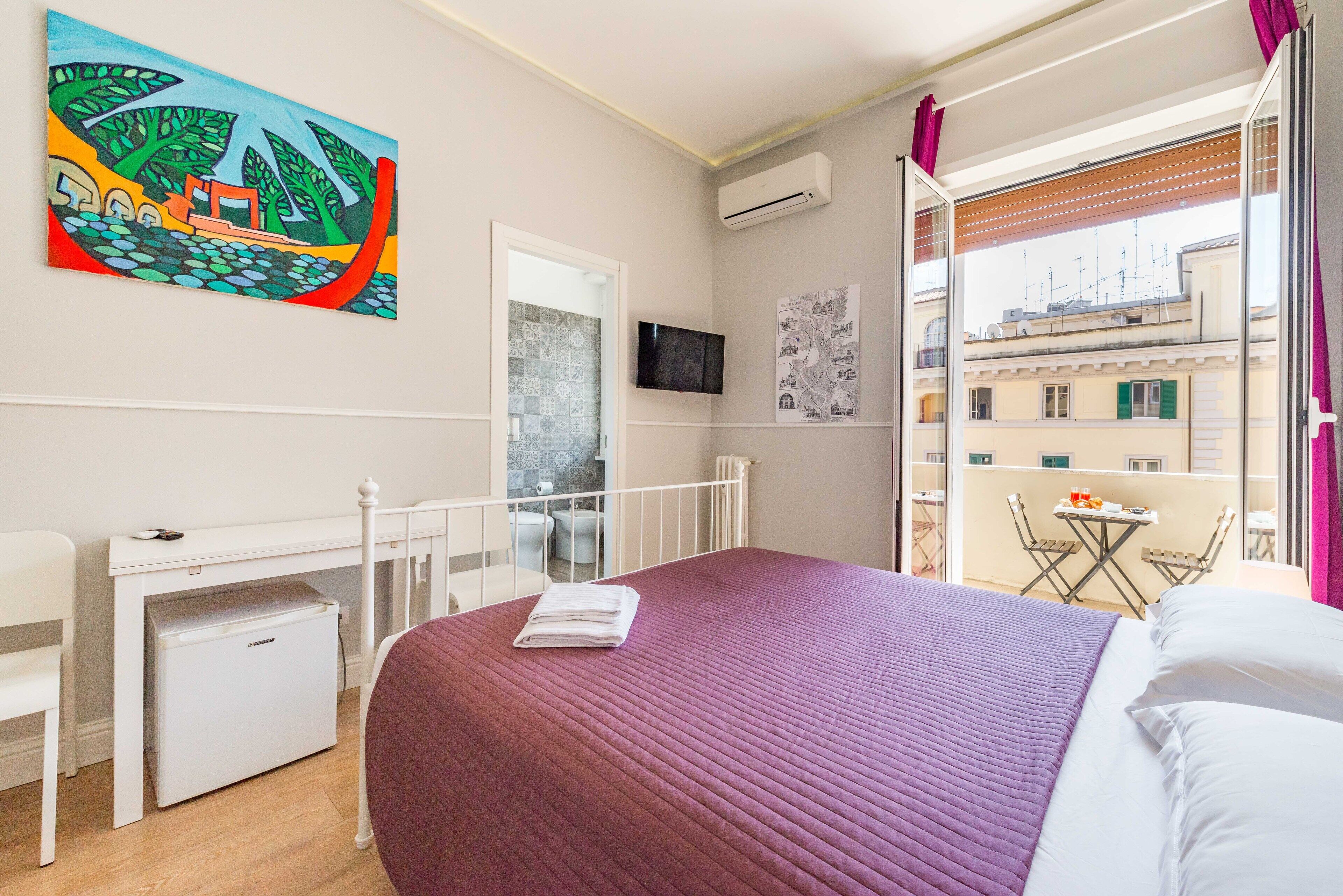 A hotel room with a bed, a desk, a small refrigerator, a TV, and a balcony with a table and chairs.