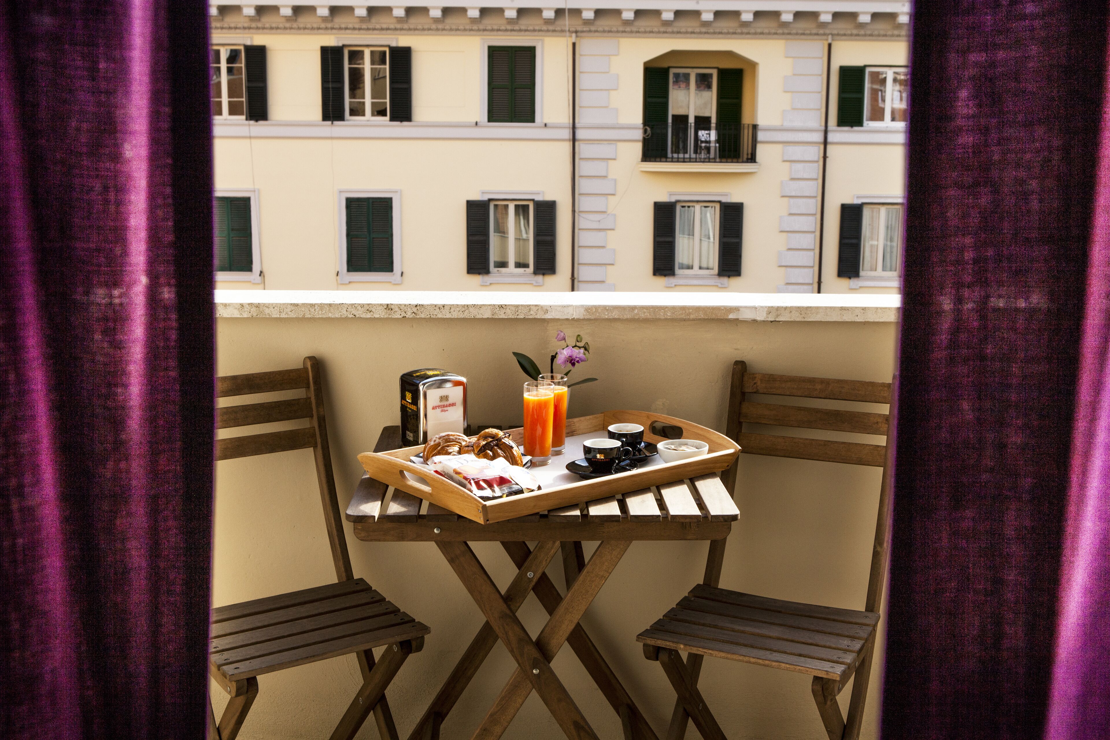 Un balcon avec une table en bois, dressée pour le petit-déjeuner, deux chaises et une vue sur des immeubles aux volets fermés.