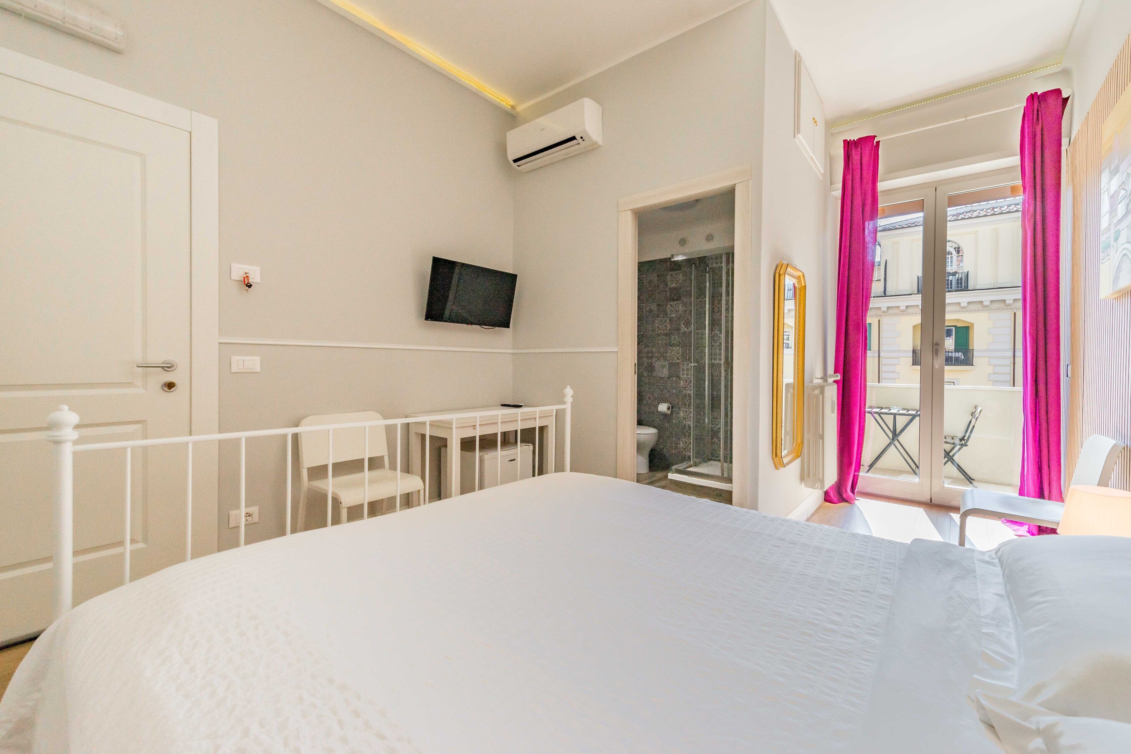Hotelkamer met een groot bed, een aan de muur bevestigde televisie, een klein bureau met stoel en een balkon met roze gordijnen.
