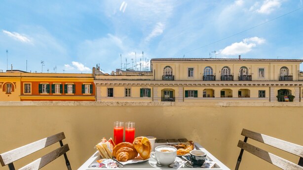 Un balcón con una mesa dispuesta para el desayuno, con cruasanes, café y jugo, con vistas a un edificio con ventanas y balcones arqueados.