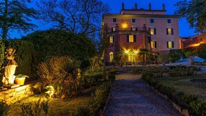 A well-lit building with a garden path leading to the entrance, surrounded by greenery and a statue.