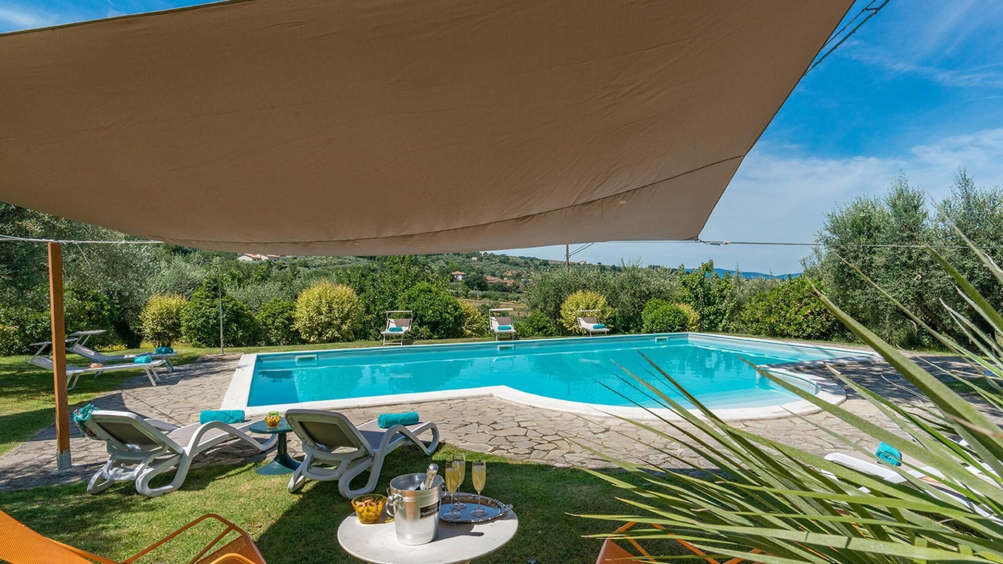 Ein Pool mit Liegestühlen und einem Tisch mit Getränken, umgeben von Grün und mit Blick auf Häuser im Hintergrund.