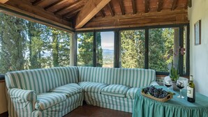 A cozy corner with a striped sofa, a table with a grape basket and wine glasses, and a view of trees through large windows. - Ar-a390-arez0at - Villalta 18 2 (Arezzo)