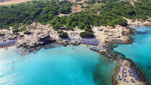 Vista aérea de una zona costera con aguas azul turquesa, costas rocosas y una playa con numerosos sombrillas azules.