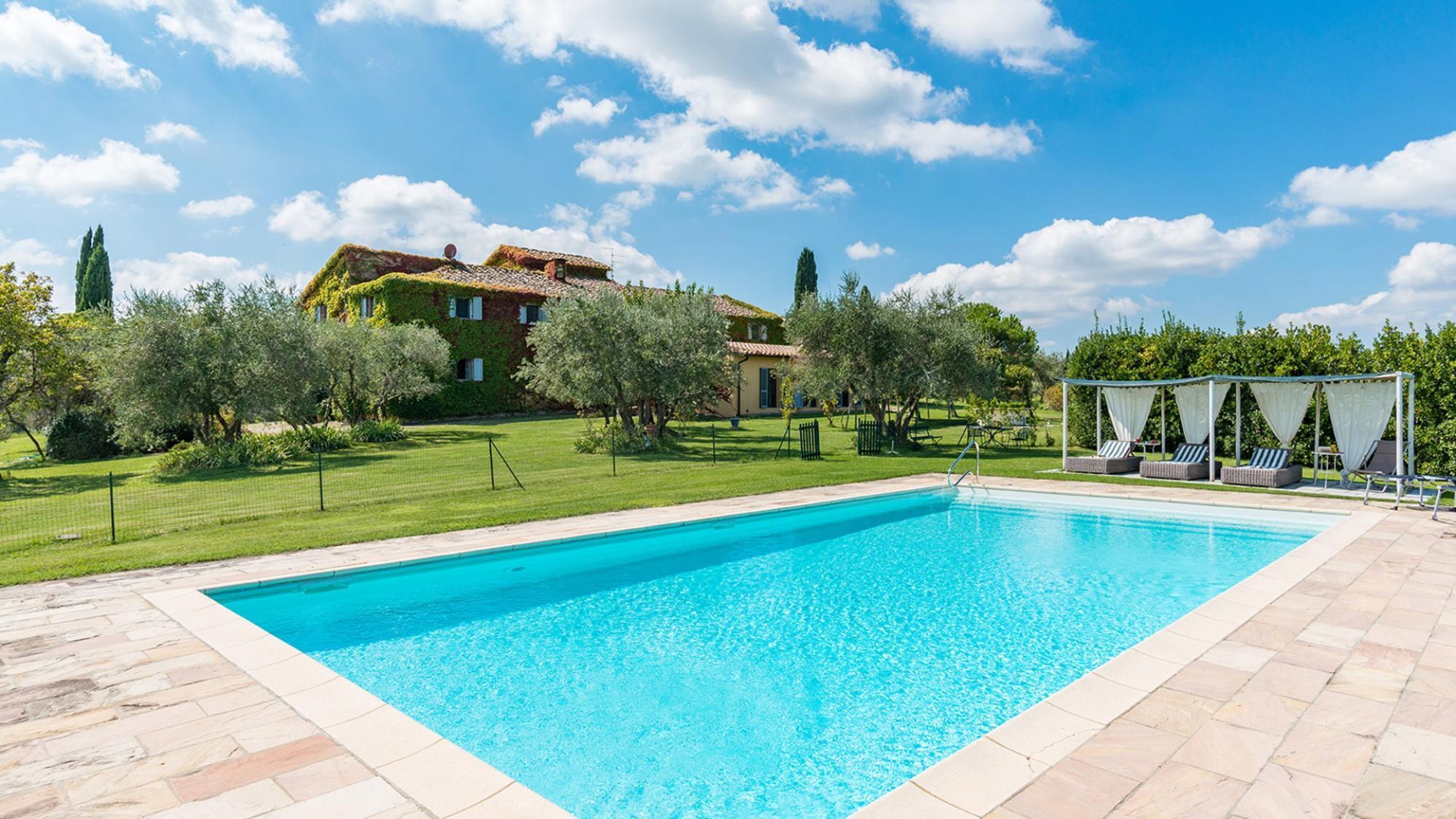 A rectangular swimming pool with a clear blue water surface, surrounded by a stone deck. In the background, there is a large house with a red-tiled roof and a garden with various trees and shrubs.