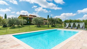 A rectangular swimming pool with a clear blue water surface, surrounded by a stone deck. In the background, there is a large house with a red-tiled roof and a garden with various trees and shrubs. - Fi-m408-snoc0at - Casabianca 12 (Barberino Tavarnelle)
