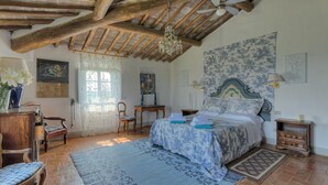A bedroom with a wooden ceiling, a bed with a floral bedspread, a chandelier, a window with lace curtains, and a wall with a floral wallpaper. - Fi-m408-snoc0at - Casabianca 12 (Barberino Tavarnelle)