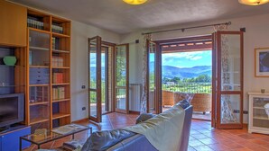 A living room with a sofa, a TV, and bookshelves. There is a glass door leading to a balcony with a view of the countryside. - Tr-f457-mcas0as - Colle del Sole 7 1 (Montecastrilli)