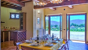 A dining area with a table set for six, a kitchen with wooden cabinets, and a view of a mountainous landscape through open doors. - Tr-f457-mcas0as - Colle del Sole 7 1 (Montecastrilli)