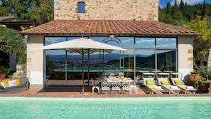 Zona piscina con tavolo e sedie, un ombrellone grande e vista su un paesaggio montano attraverso ampie finestre.
