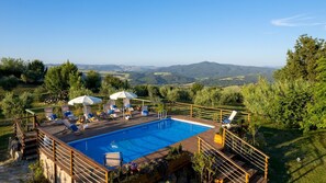 Zona de piscina con sillas de descanso y sombrillas, rodeada por una terraza de madera y con vistas a un paisaje pintoresco.