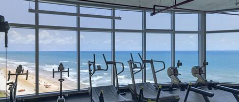A gym with large windows overlooking a beach, featuring treadmills, elliptical machines, and weight benches.