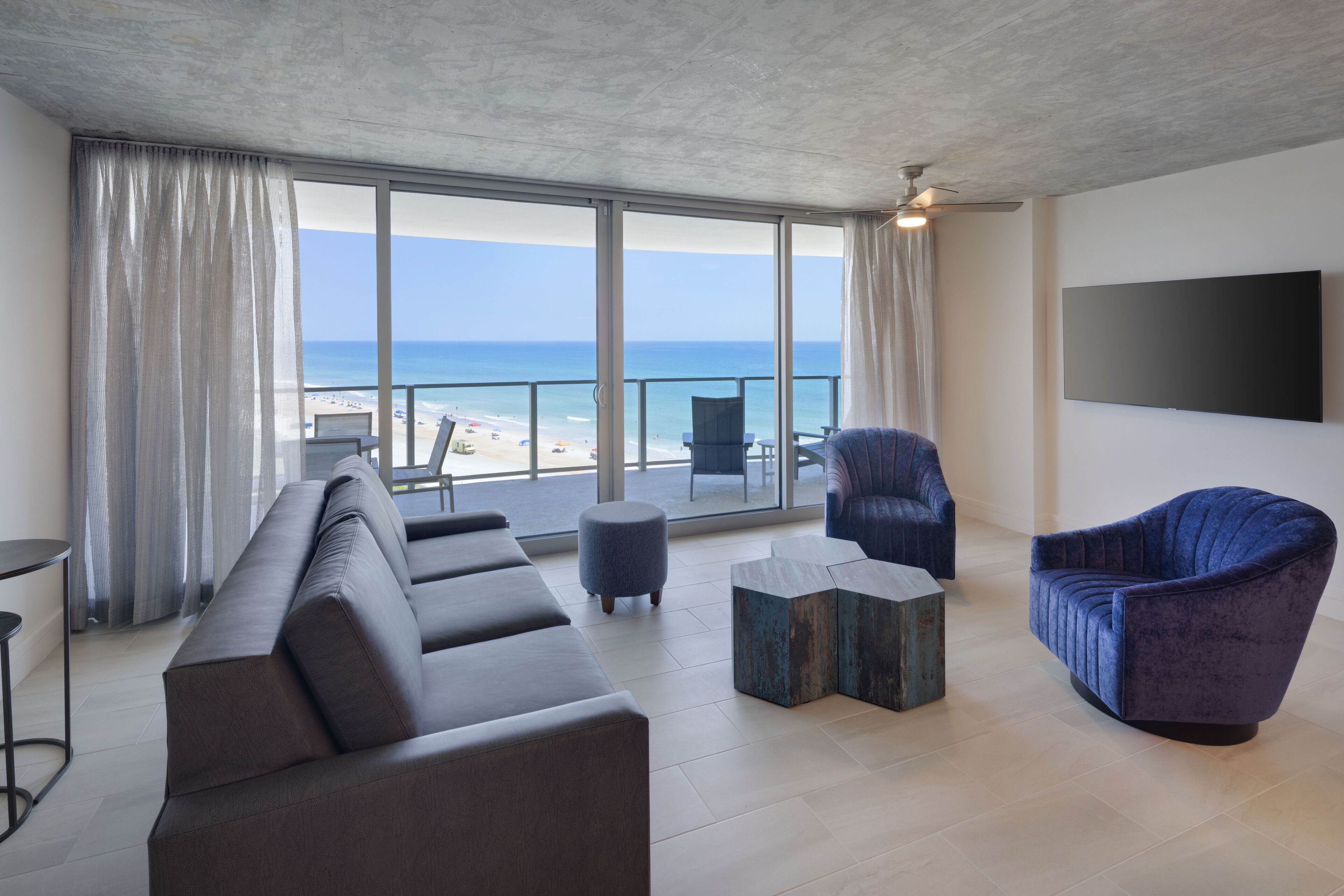 A modern living room with a large window overlooking a beach, a grey sofa, a blue armchair, and a flat-screen TV on the wall.