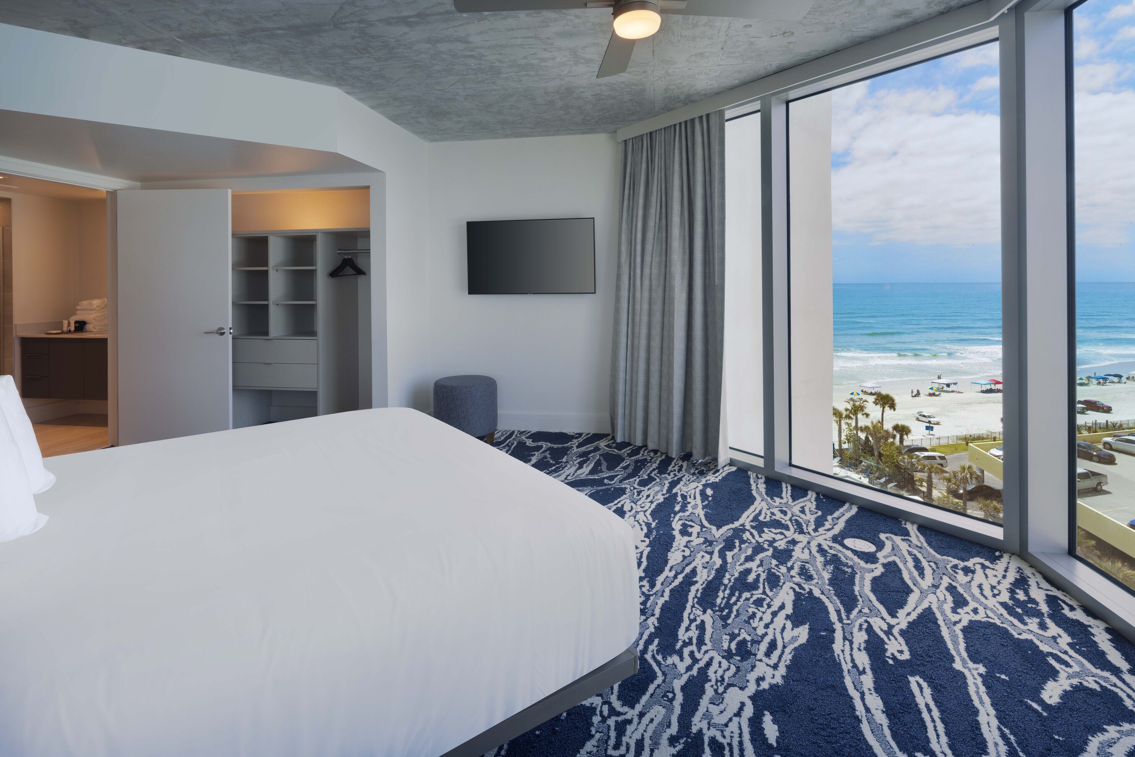 A hotel room with a large bed, a flat-screen TV, a view of the beach, and a walk-in closet.