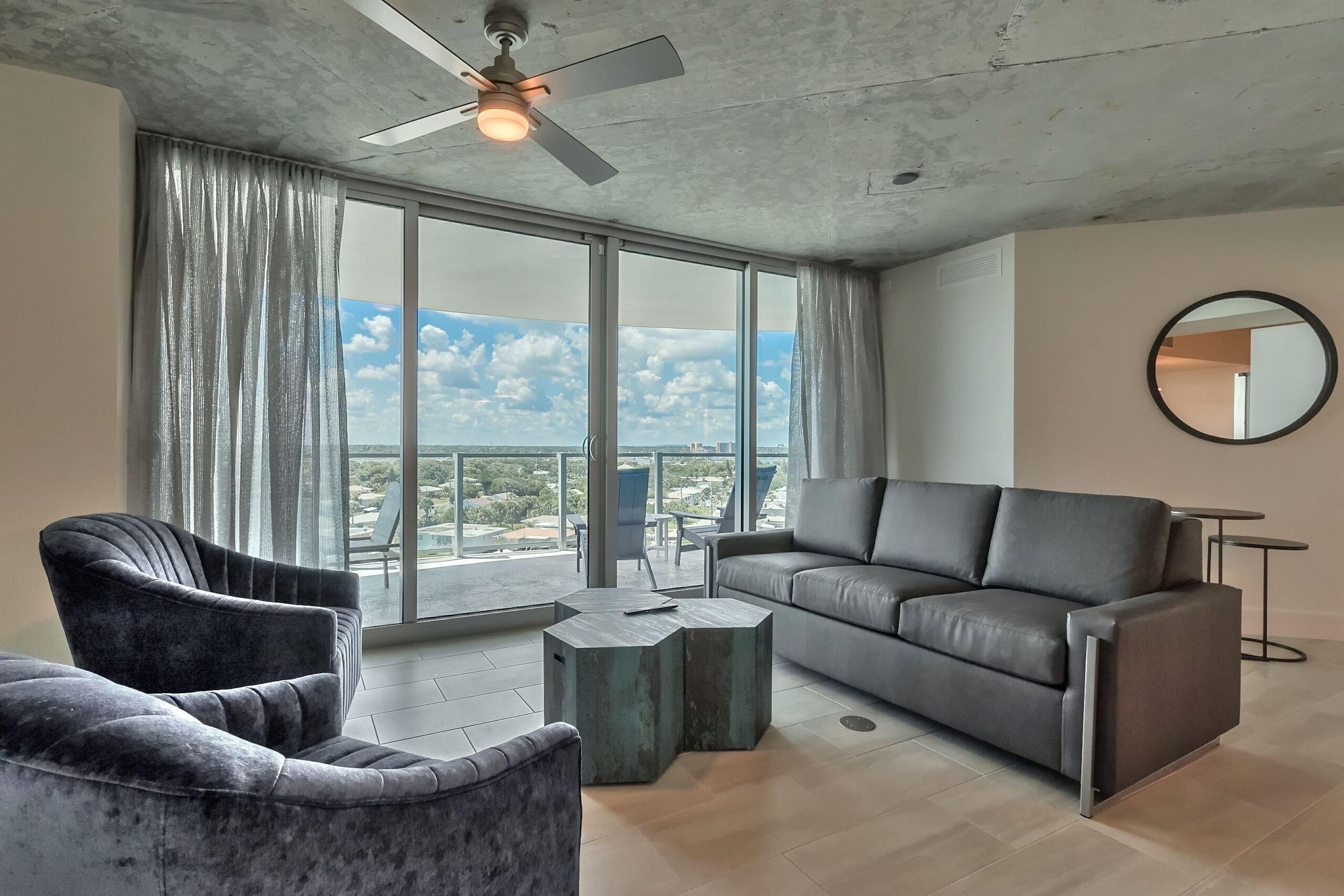 A modern living room with a sofa, armchair, and coffee table, large windows with curtains, a ceiling fan, and a round mirror on the wall.