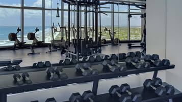 A gym with various exercise equipment and dumbbells arranged on racks, with a large window overlooking a beach and ocean view.