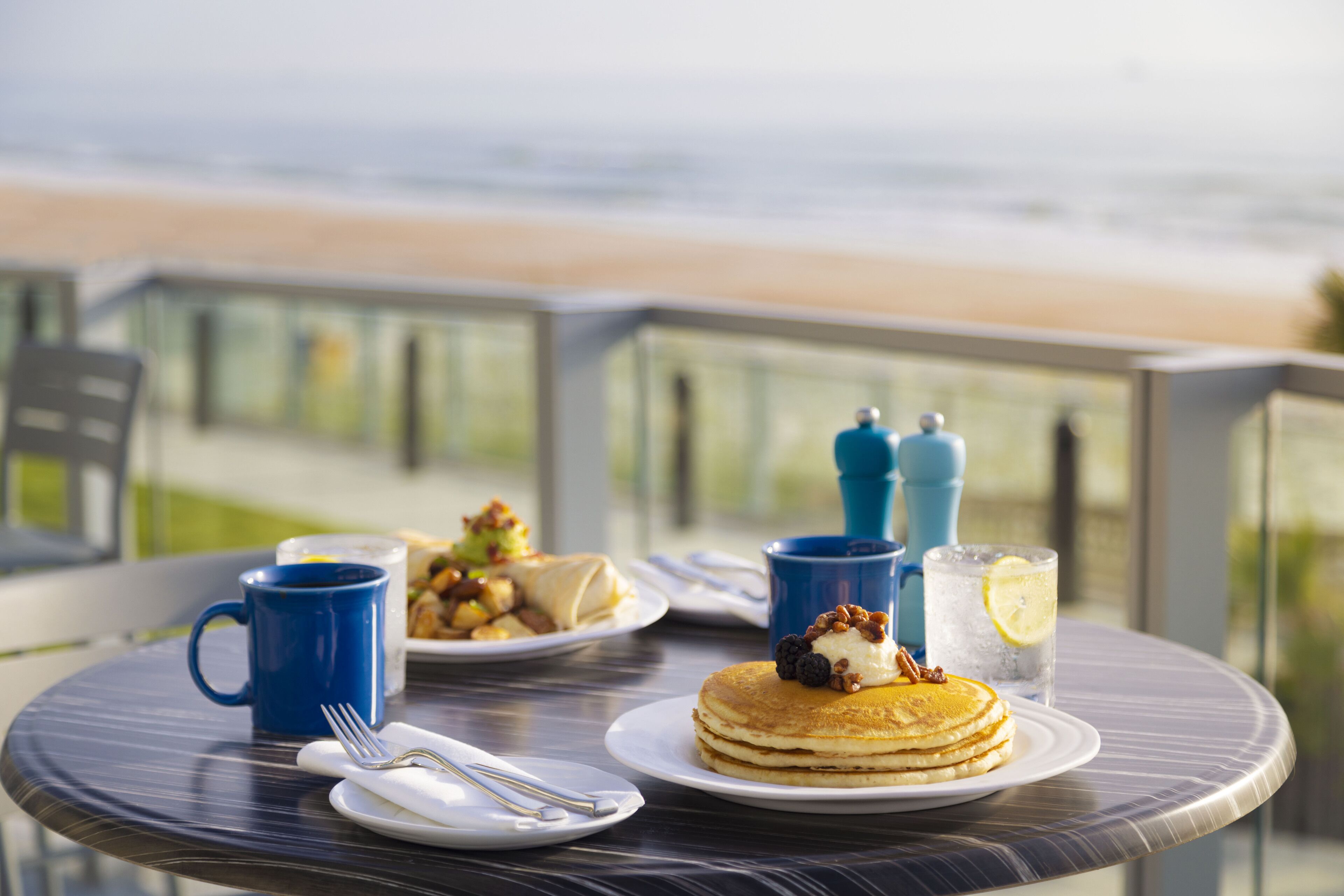 A breakfast table with pancakes, a mug, and a glass of iced tea on a balcony overlooking the beach.