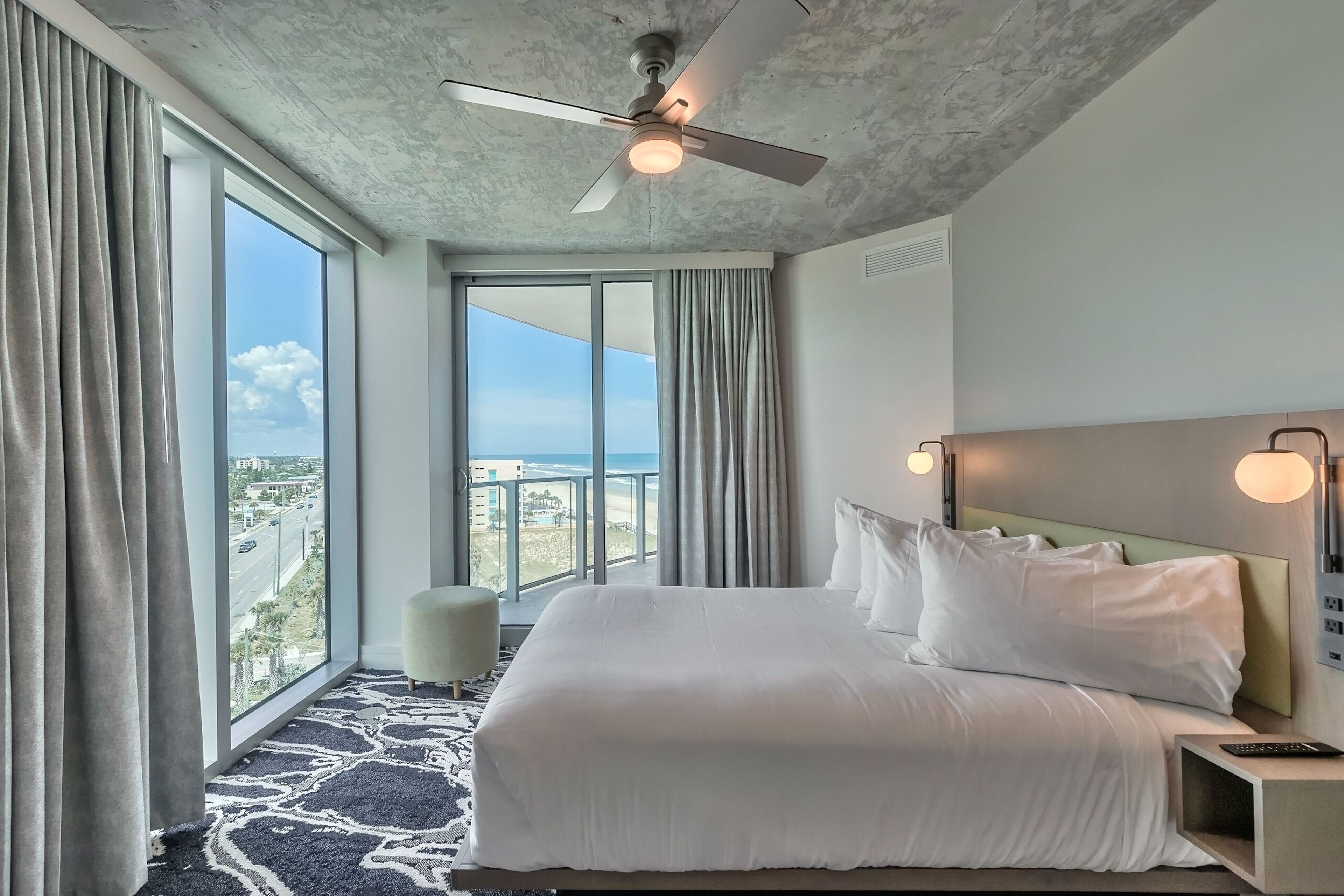 A hotel room with a large bed, a ceiling fan, a view of the city, and a balcony.