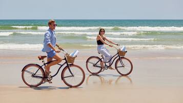 Duas pessoas andando de bicicleta na praia, com o oceano ao fundo.