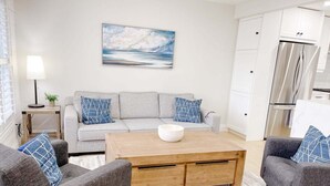 Ein modernes Wohnzimmer mit einem grauen Sofa, passenden Sesseln, einem Holztisch und einem Gemälde, das eine Meereslandschaft zeigt.