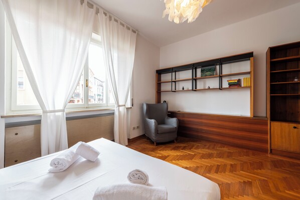 A hotel room with a bed, a chair, a wooden floor, a bookshelf, and a window with curtains. - Riverview Luxury Flat (Verona)