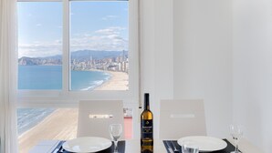 A dining table with two plates and wine glasses, a bottle of wine, and a view of a beach and cityscape through a window. - Brilliant Panoramic Front line apartment Veracruz Levante (R099) (Benidorm)