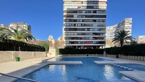 A swimming pool with clear blue water, surrounded by a low brick wall and a fence. In the background, there are tall residential buildings. - Brilliant Panoramic Front line apartment Veracruz Levante (R099) (Benidorm)