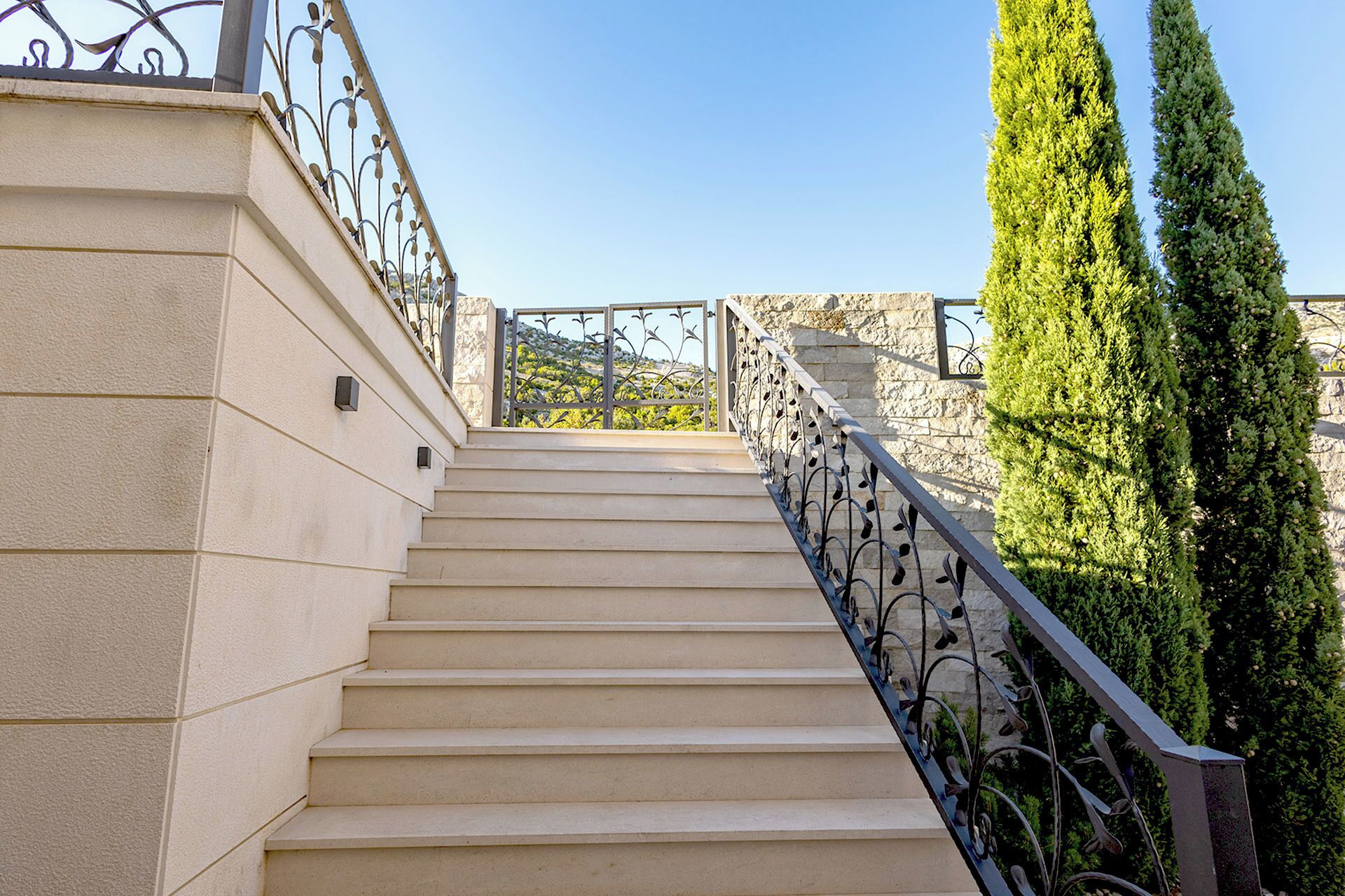 Un escalier aux marches de couleur beige et aux rampes en métal noir, menant à un mur de pierre d’où l’on peut admirer un paysage verdoyant et des collines au loin.