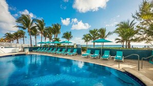 A pool area with lounge chairs and umbrellas, palm trees, and a view of the ocean. - Private Beachfront Condominium with Resort and Amenities Access (Sunny Isles Beach)