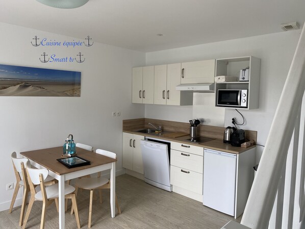 A modern kitchen with white cabinetry, a wooden countertop, a dishwasher, and a microwave. There is a dining table with chairs and a picture of a beach on the wall.