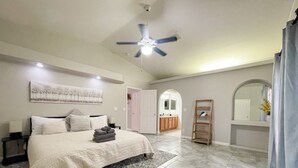 Quarto espaçoso com uma cama grande, um ventilador de teto e um banheiro visível por um arco.