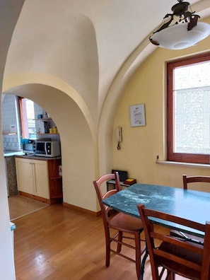 A dining area with a table and chairs, a kitchen in the background, and a window with patterned curtains.