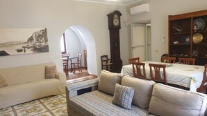 Ein Wohnzimmer mit einer Couch, einem Esstisch mit Stühlen, einer Uhr und einem Bücherregal.