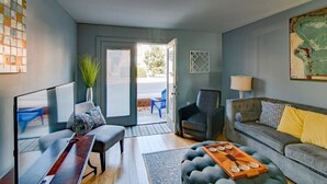 A modern living room with a grey sofa, a coffee table, and a TV. There is a door leading to an outdoor area with a blue chair and a plant.