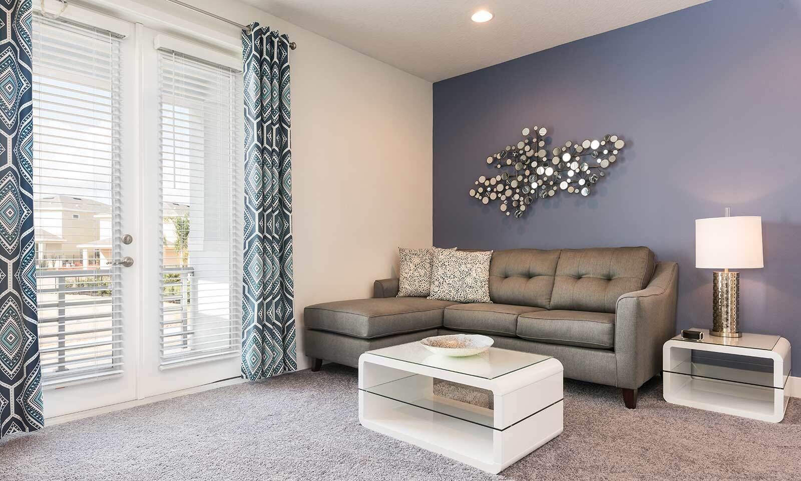 A living room with a sectional sofa, a glass coffee table, a side table with a lamp, and a decorative wall piece.