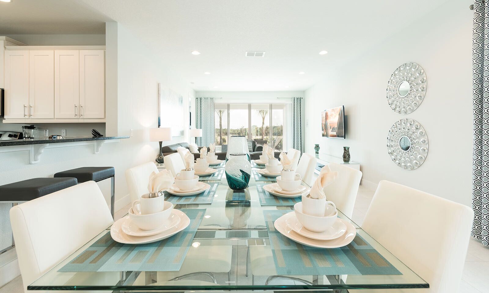 A modern dining area with a glass table set for a meal, white chairs, and a kitchen in the background.
