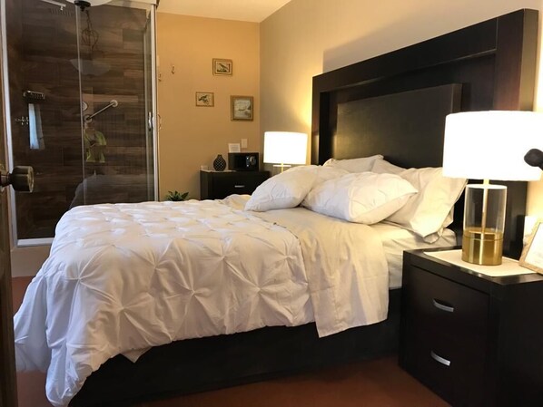 A hotel room with a bed, a nightstand, a lamp, a glass shower enclosure, and a wall with framed pictures.