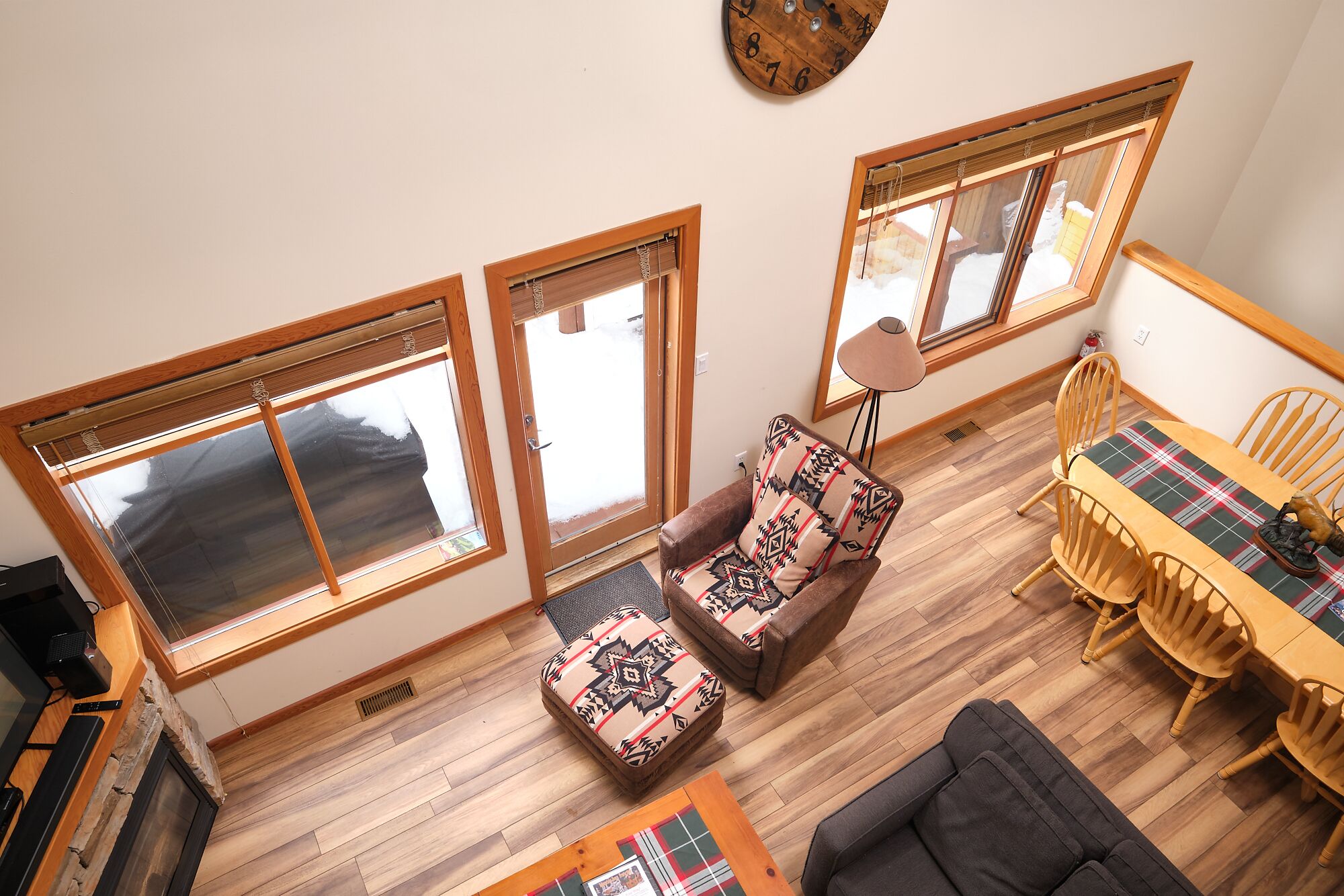 Un salon chaleureux avec un plancher en bois, un canapé agrémenté de coussins à motifs, une table à manger avec des chaises et de grandes fenêtres équipées de stores.