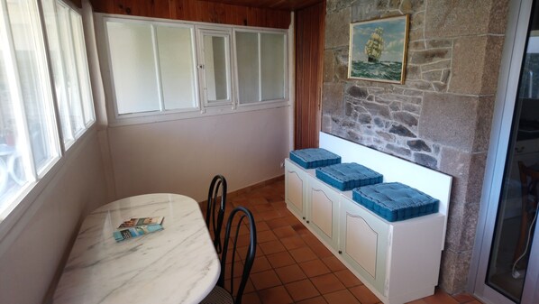 Una habitación pequeña con una mesa blanca, sillas, un armario con cajas azules y una ventana, junto a una pared de piedra con un cuadro enmarcado.