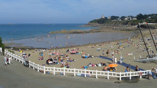 A crowded beach with people sunbathing and swimming, a white fence, and a lighthouse in the background.