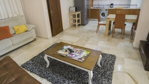 Un espace de vie compact comprenant un canapé, une table basse avec des magazines, un lave-linge et un petit coin cuisine.
