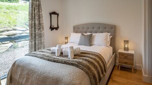A neatly made bed with a grey headboard, white linens, and a brown throw blanket. There are two bedside tables with lamps, a mirror on the wall, and a window with curtains. - 6 Loch View With Loch Views (Aberfeldy)
