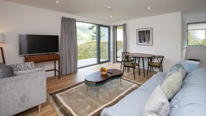 A modern living room with a sofa, a coffee table, a TV, a dining table with chairs, and a large window with curtains. - 6 Loch View With Loch Views (Aberfeldy)