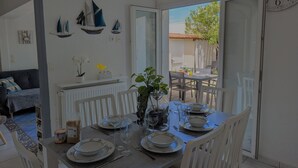 A dining area with a table set for four, white chairs, and a view of the outdoors through a glass door. - Sol-y-Days Grau Village, Holiday home with large terrace (Le Grau-du-Roi)